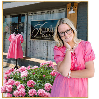 Kendra Kay's Boutique. Woman in pink dress standing in front of a boutique storefront with pink flowers and mannequin our front. 